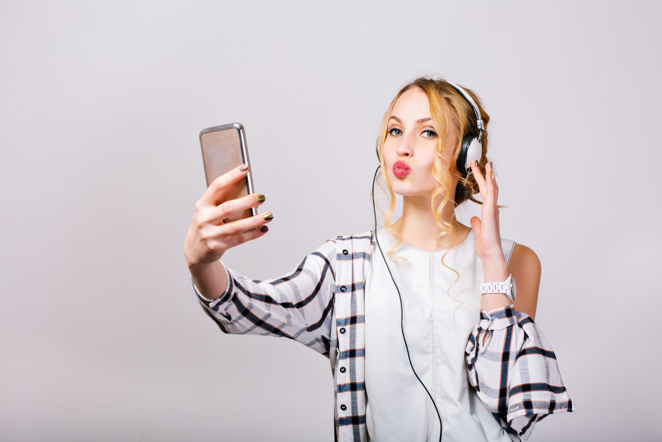 Gorgeous Cute Young Woman Taking Selfie Near Grey Wall Looking Smartphone Sending Kiss Mirthful Blonde Girl Wearing Plaid Shirt White Blouse Happy Brightful Emotions Isolated 1 Scaled